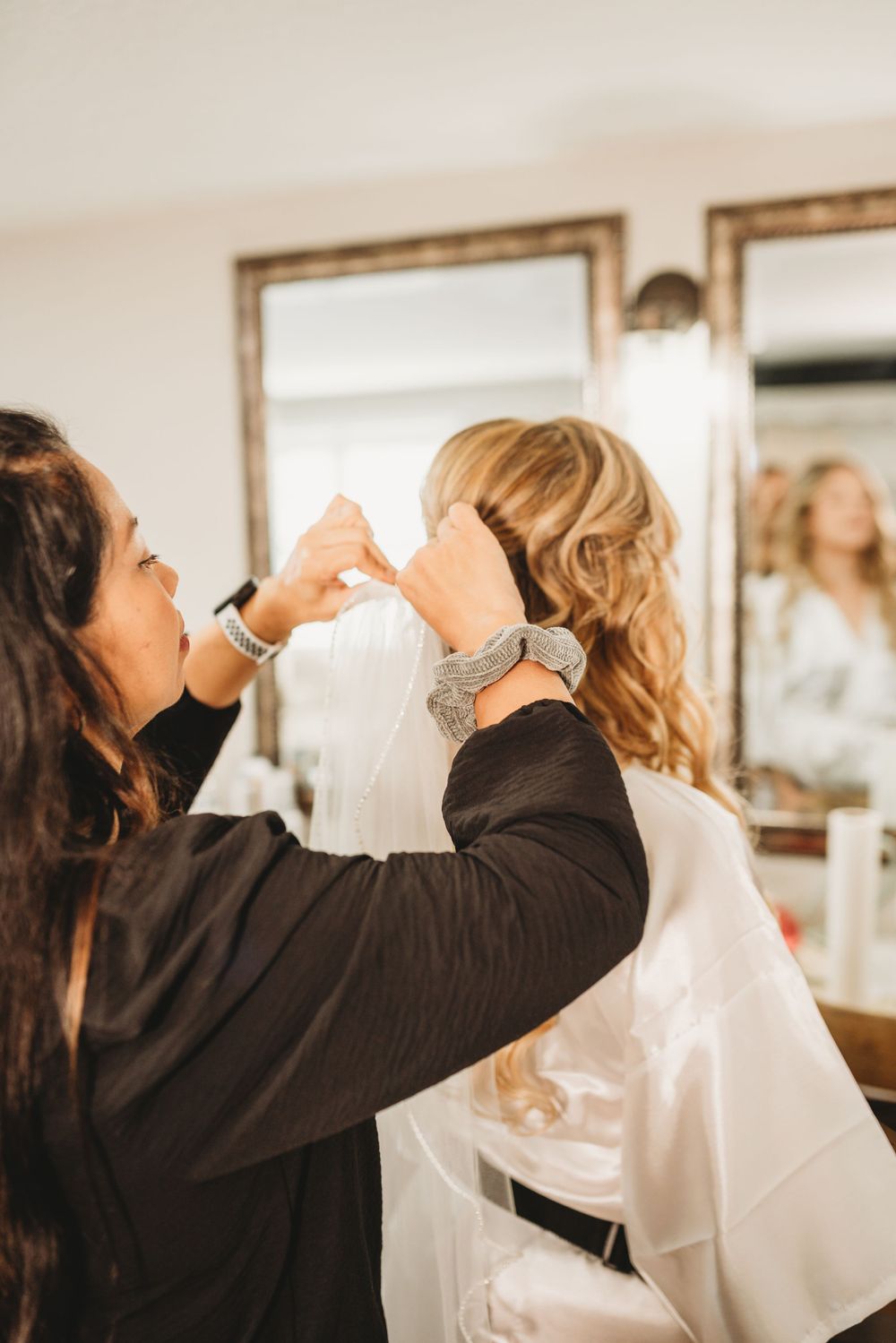 Stylist putting finishing touches on bridal hair preparation in front of vanity mirror.