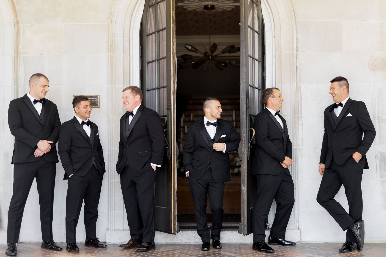 Groomsmen in black tuxedos pose playfully in sequence along white building exterior.