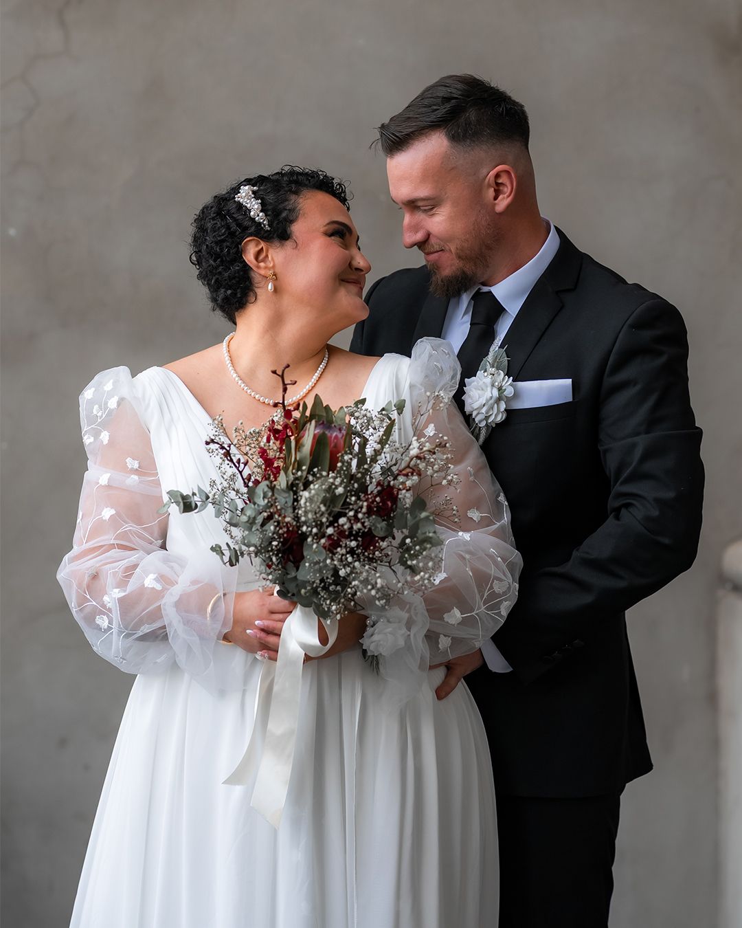 Melbourne Bride and groom gazing at each other, she holds a red and white bouquet, wearing a white gown, he in a black suit.