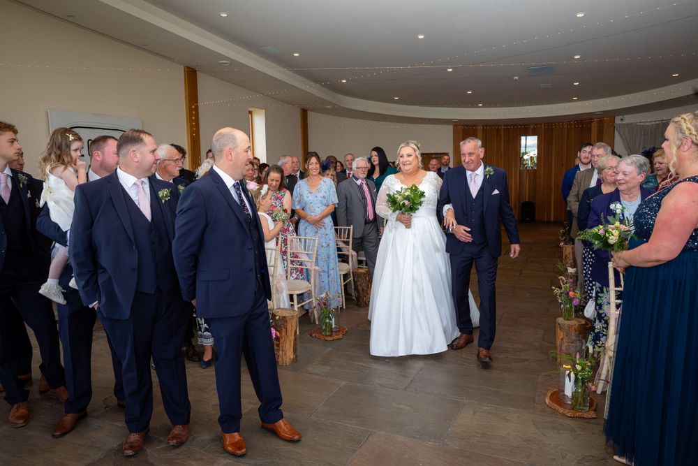 Bride walks down the aisle with her groom through applauding guests in a bright modern wedding ceremony venue.