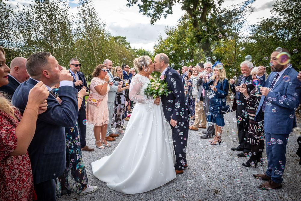 Newlyweds share a tender kiss surrounded by guests blowing bubbles during an outdoor wedding confetti celebration.