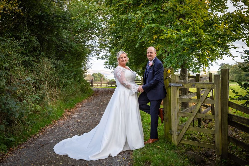 Bride and groom pose romantically by a rustic wooden gate on a scenic countryside path lined with trees.