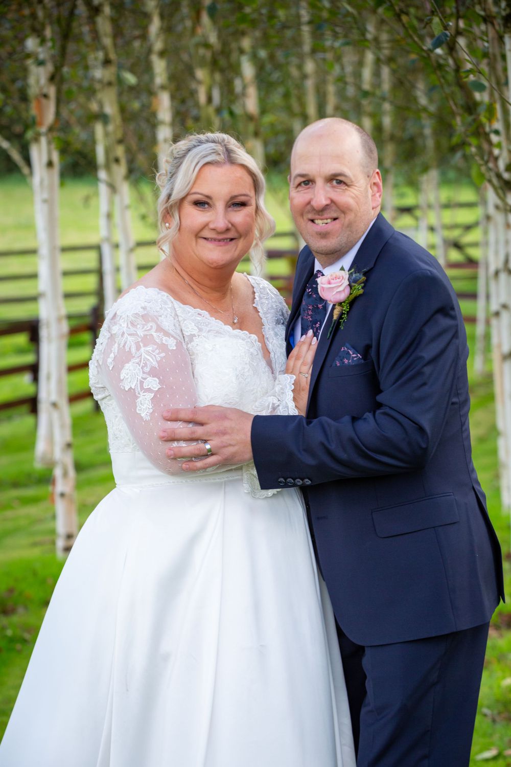 Smiling bride in lace wedding gown embraces groom in navy suit among silver birch trees in a green garden.