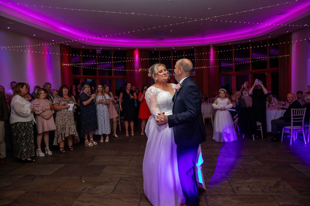 Bride and groom sharing their first dance surrounded by guests at a wedding reception with purple lighting.