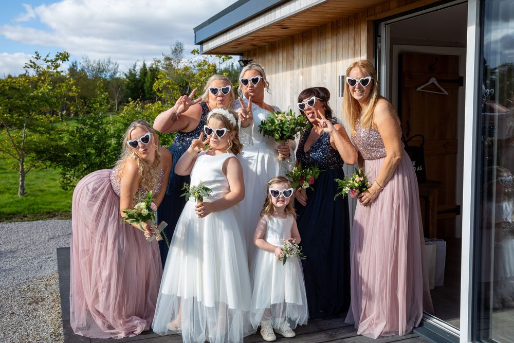 Bride with bridesmaids and flower girls in pink and navy dresses pose playfully wearing matching sunglasses outdoors.