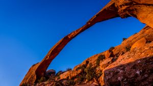 Natural stone arch formation glows orange in morning light against vibrant blue sky.