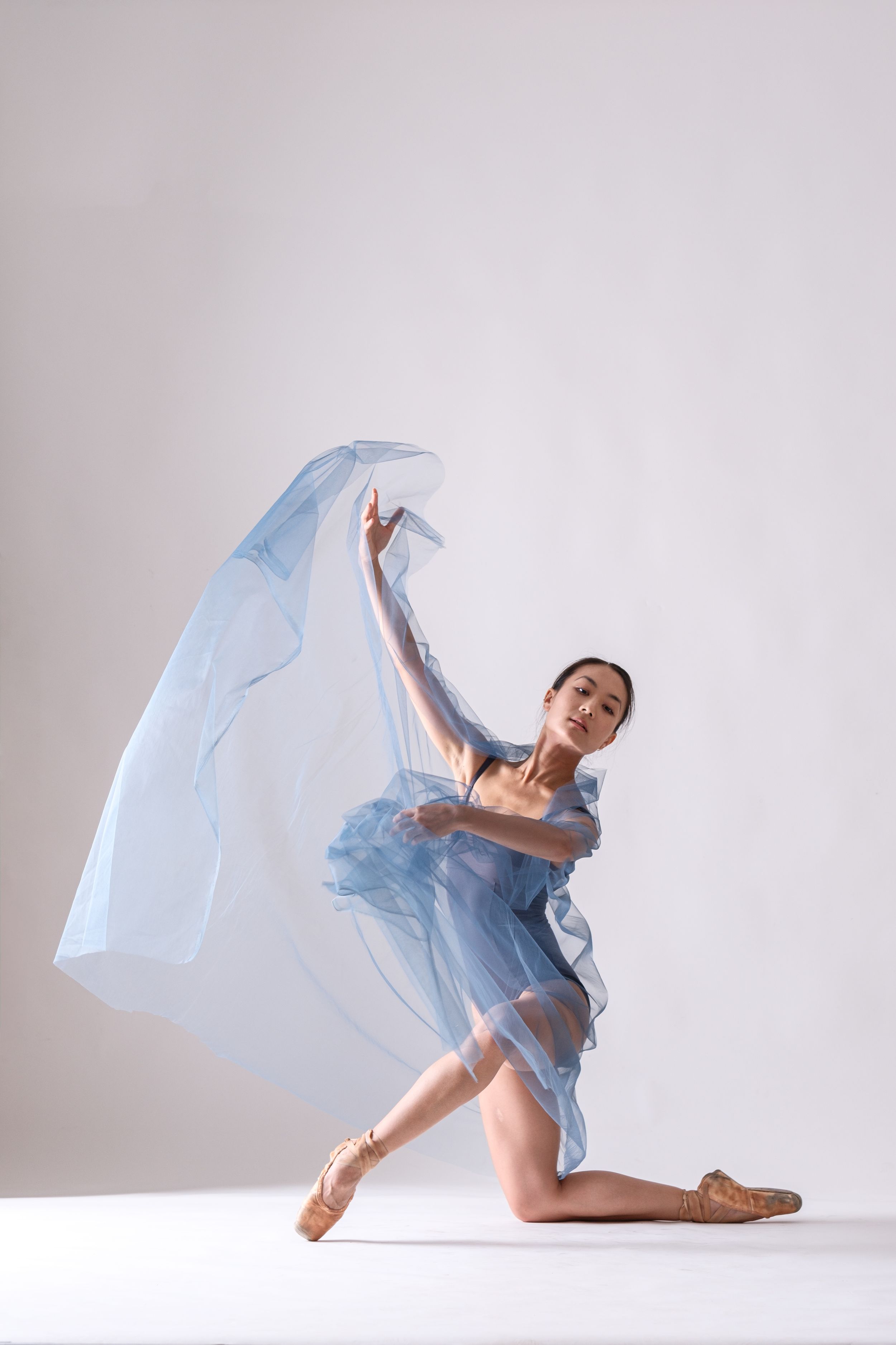 Graceful dance pose with ethereal blue fabric creating movement against white background.