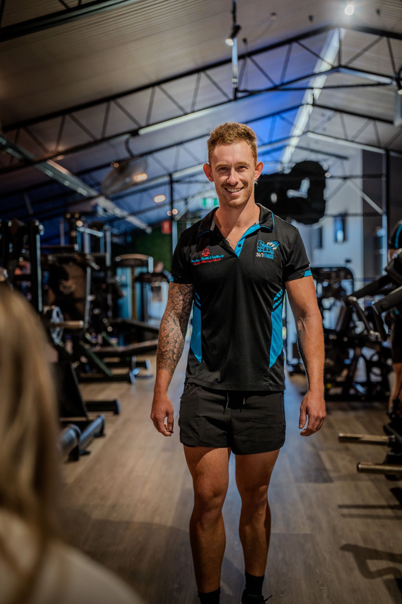 Athletic trainer in black and blue polo shirt and shorts standing in modern fitness facility with exercise equipment.