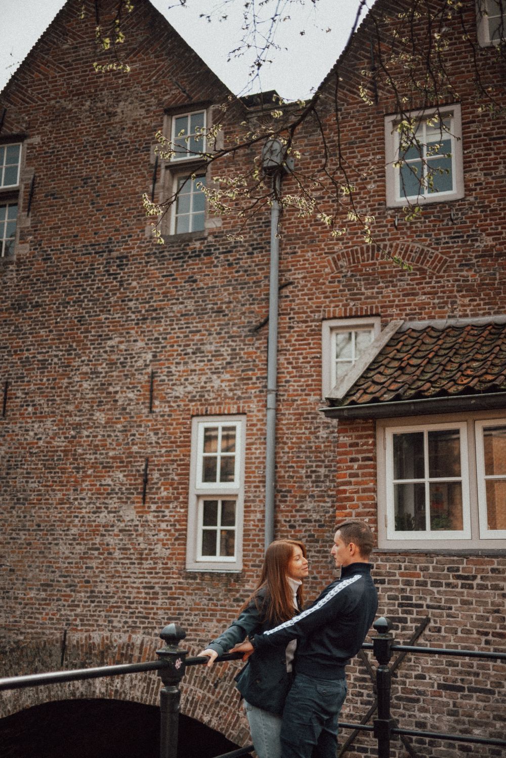 A couple walks along a historic brick building exterior with arched windows and traditional architecture.