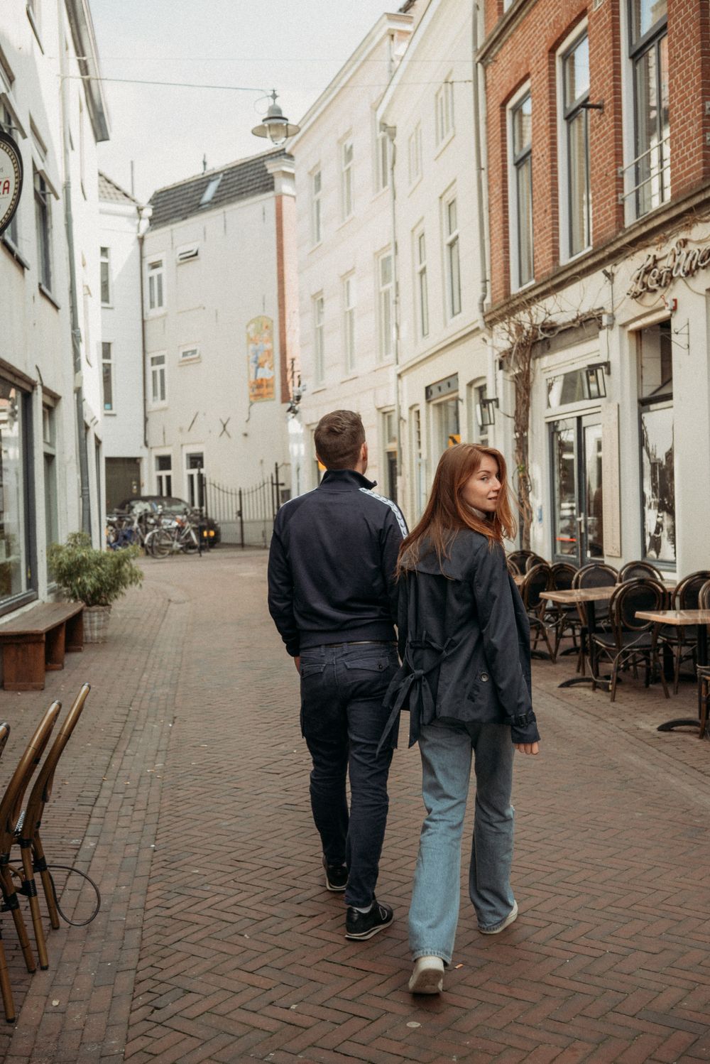 Two people stroll down a charming cobblestone street lined with brick buildings in a European town.