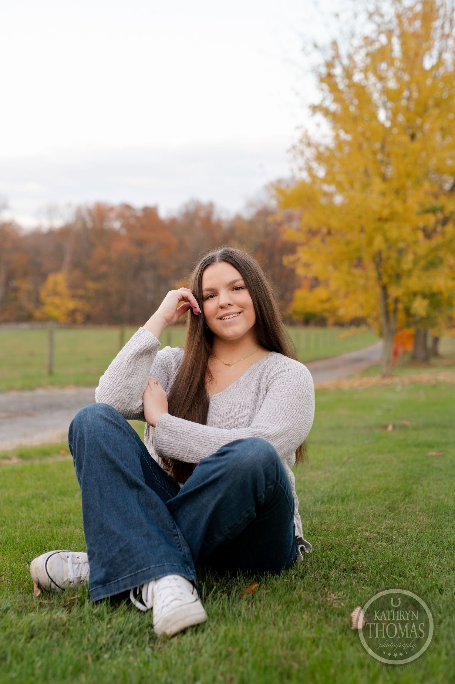 Person relaxes on grassy hill wearing casual sweater and jeans against autumn trees.