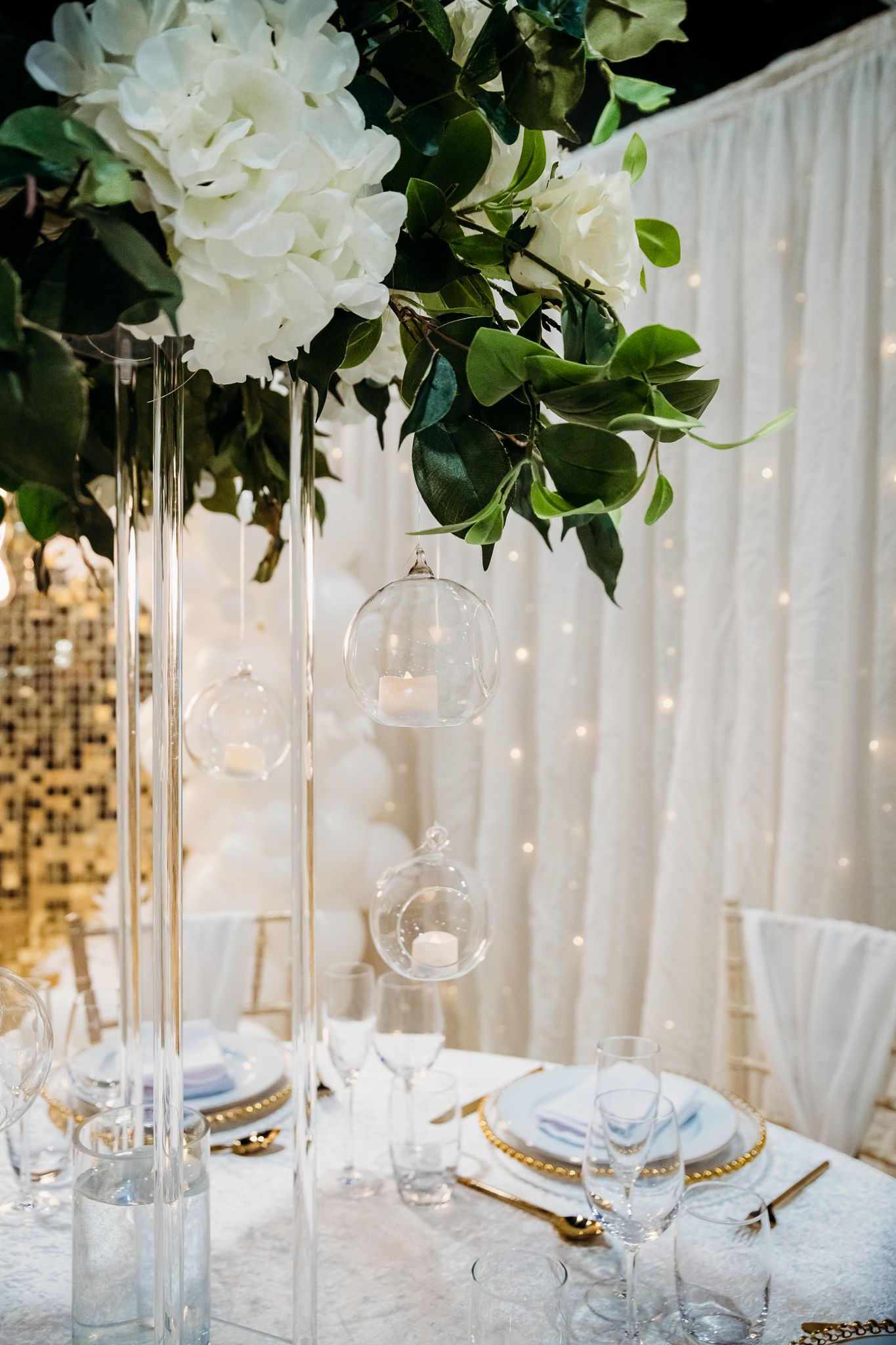 Elegant wedding table setting with tall glass vases, white hydrangeas, greenery, hanging glass orbs, and gold-rimmed charger plates.