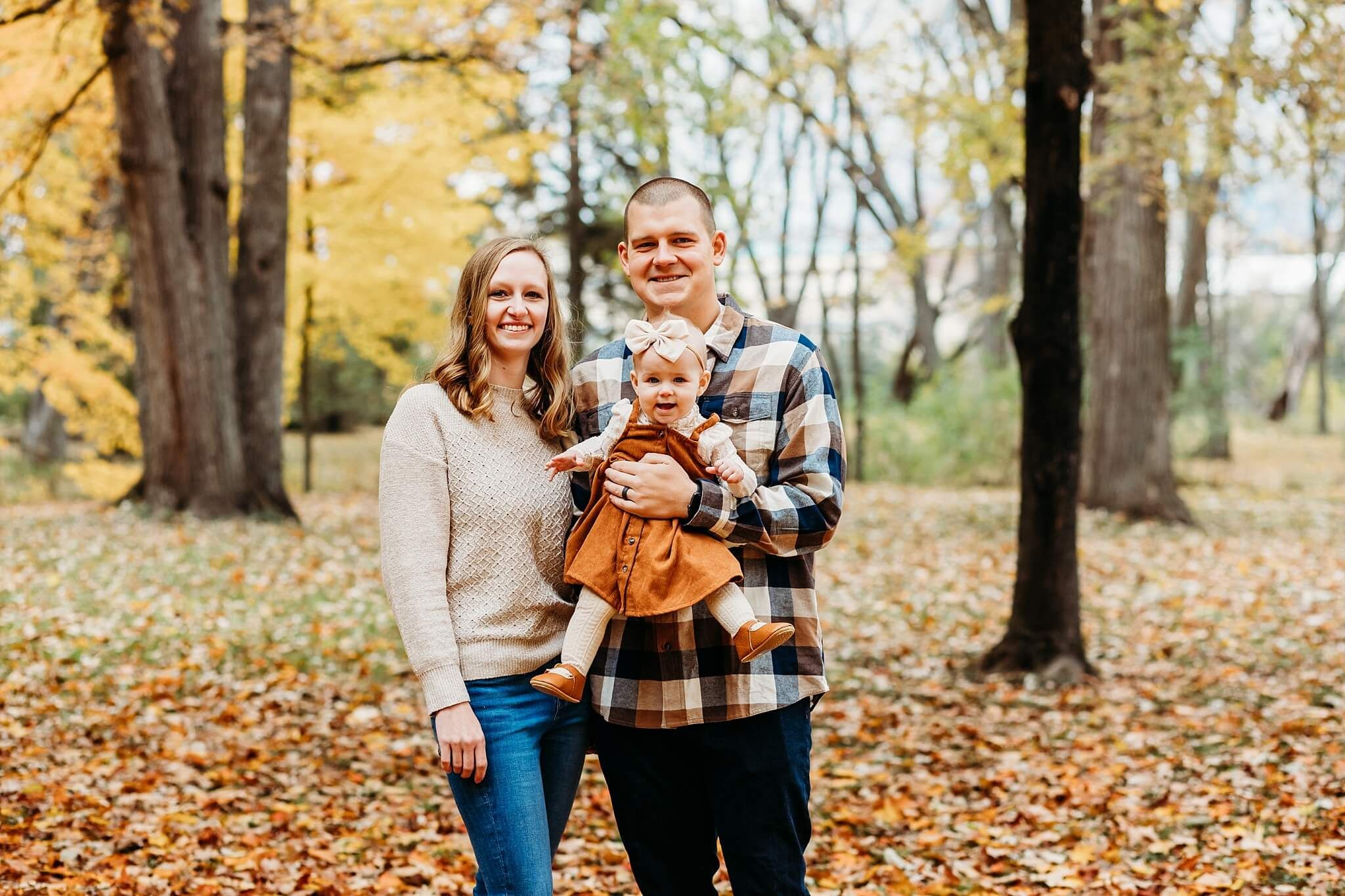 A family of three poses in a forest with fallen autumn leaves wearing cozy sweaters.