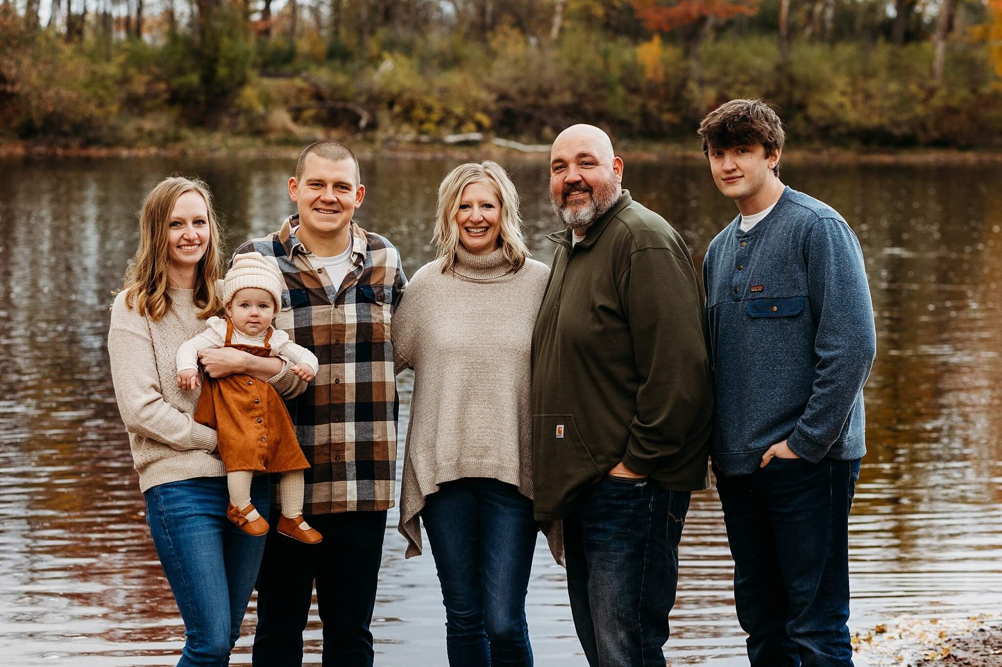 A family group poses beside a lake in autumn wearing coordinated casual outfits.