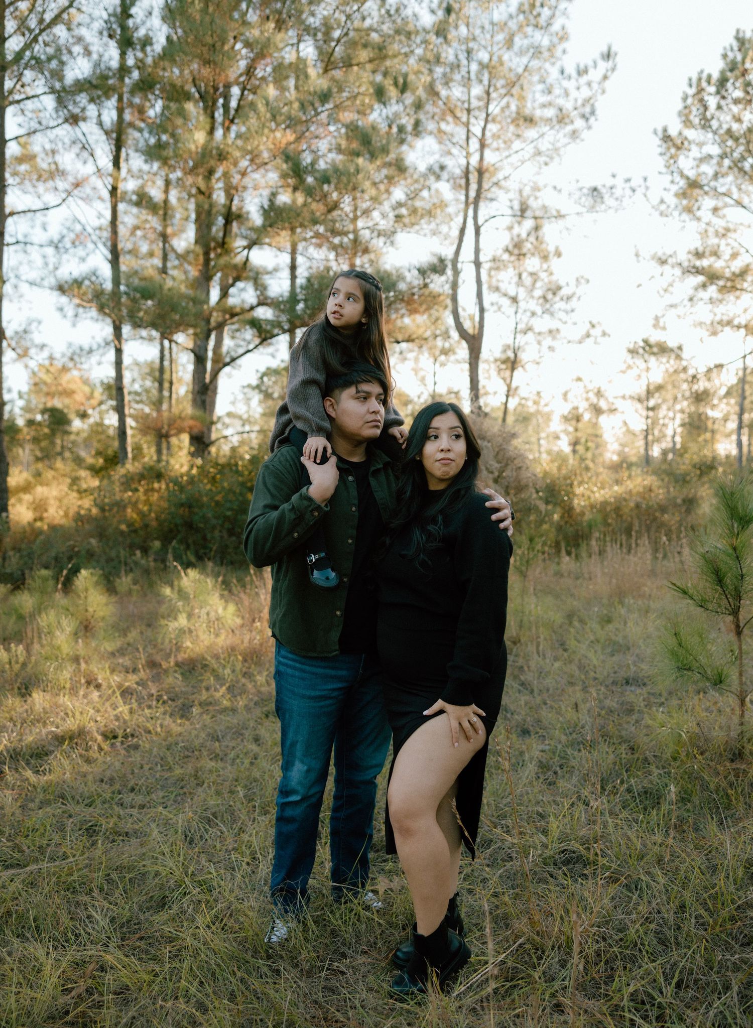 A family of three pose together in a wooded area with tall pine trees and golden sunlight filtering through the branches.