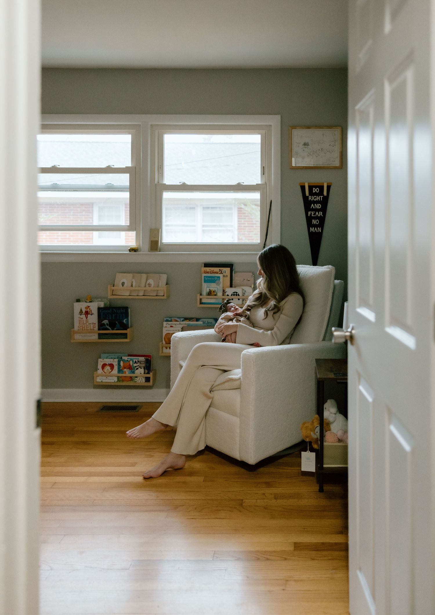 A cozy nursery corner with a white glider chair, hardwood floors, and children's books displayed on low shelves near windows.