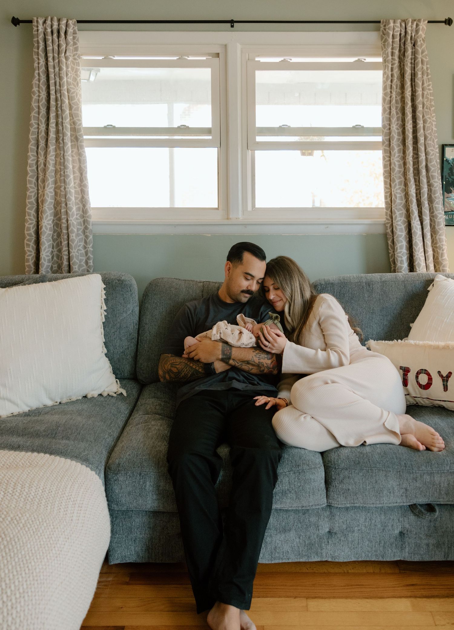 At home newborn session. A couple shares an intimate moment together while relaxing on a gray couch in a bright, sunlit room.
