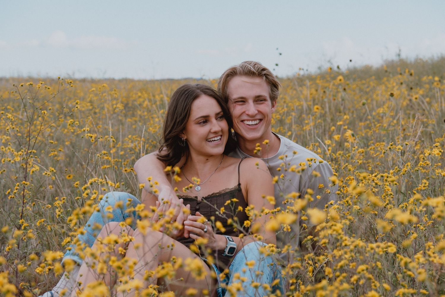 Couple poses playfully in a blooming meadow of yellow wildflowers against blue sky background.