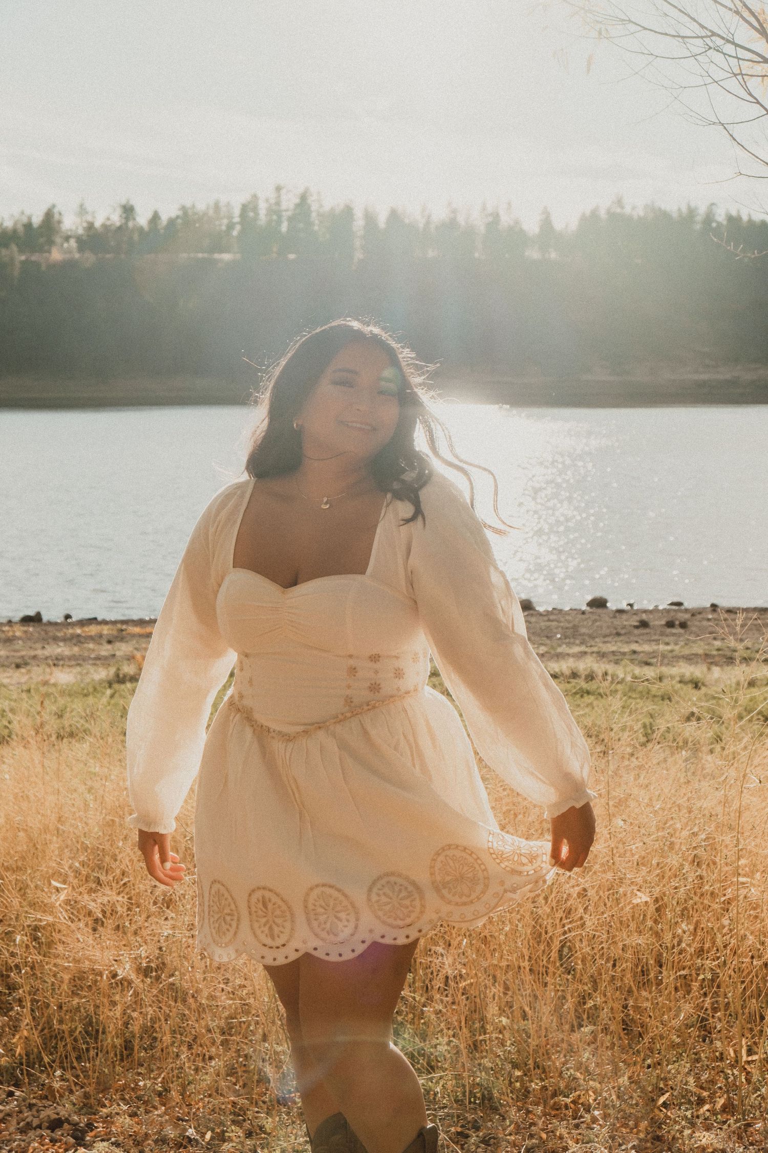 Golden hour portrait of a person in a white eyelet lace dress walking through a sun-lit field by a serene lake.