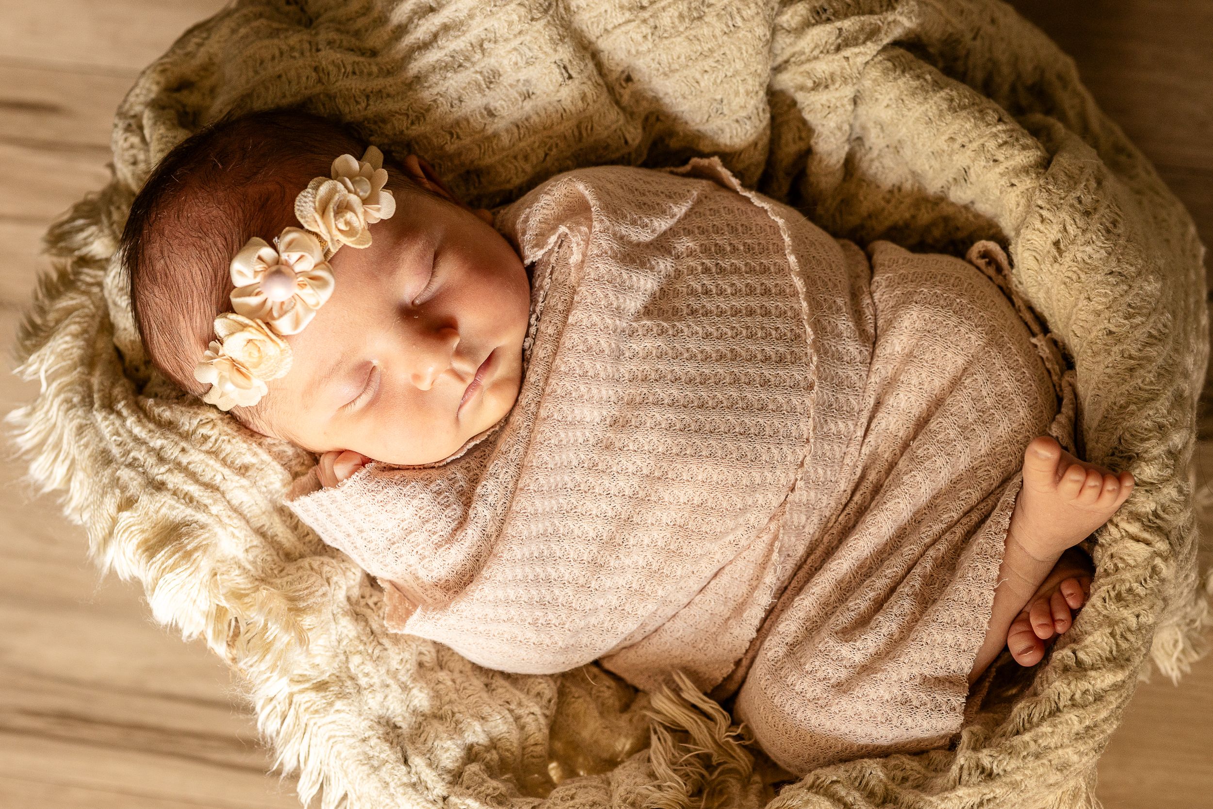 Sleeping newborn wrapped in beige swaddle lying on cozy fur blanket with delicate floral crown.