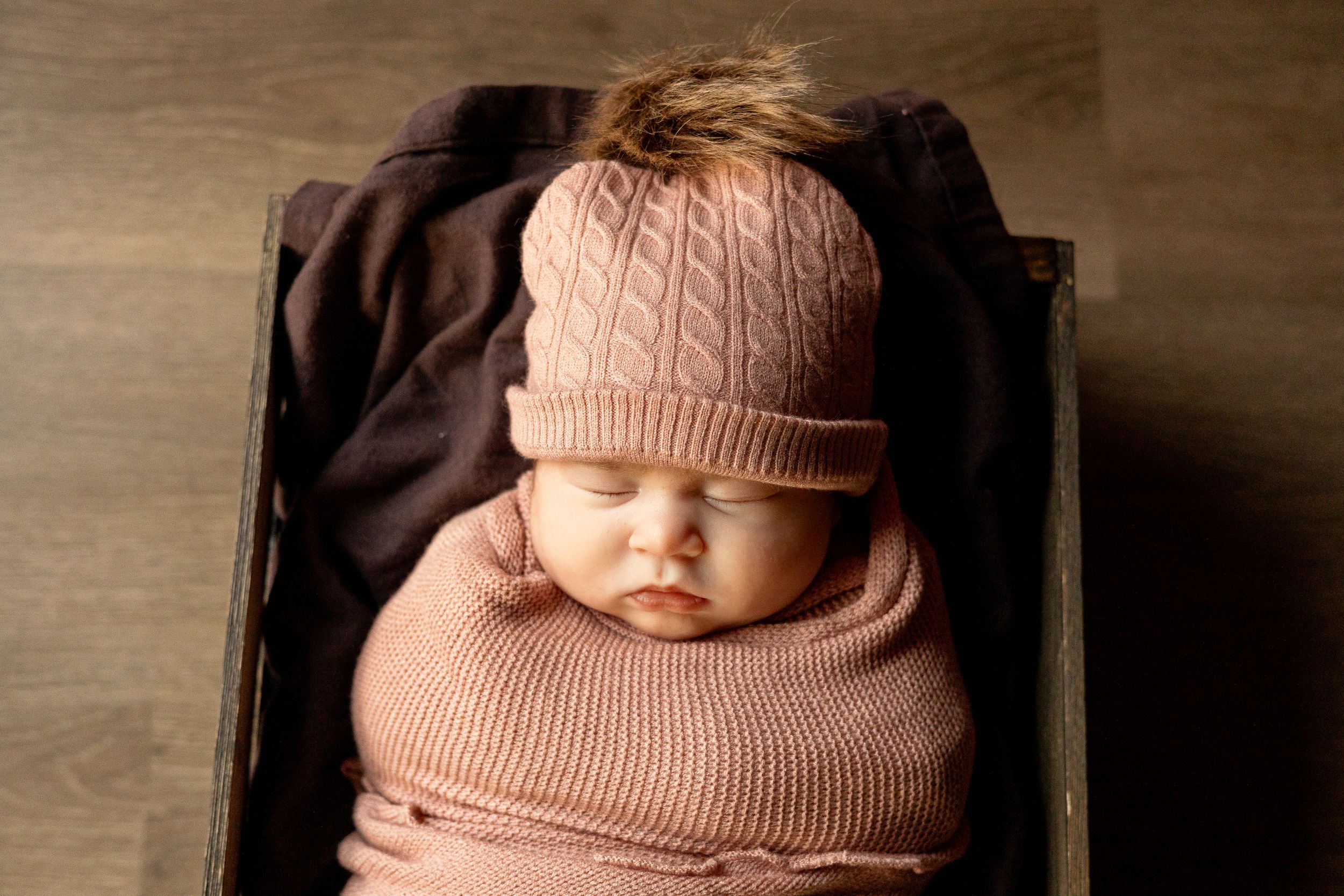 Baby wearing pink knit hat and sweater sleeping in wooden box.