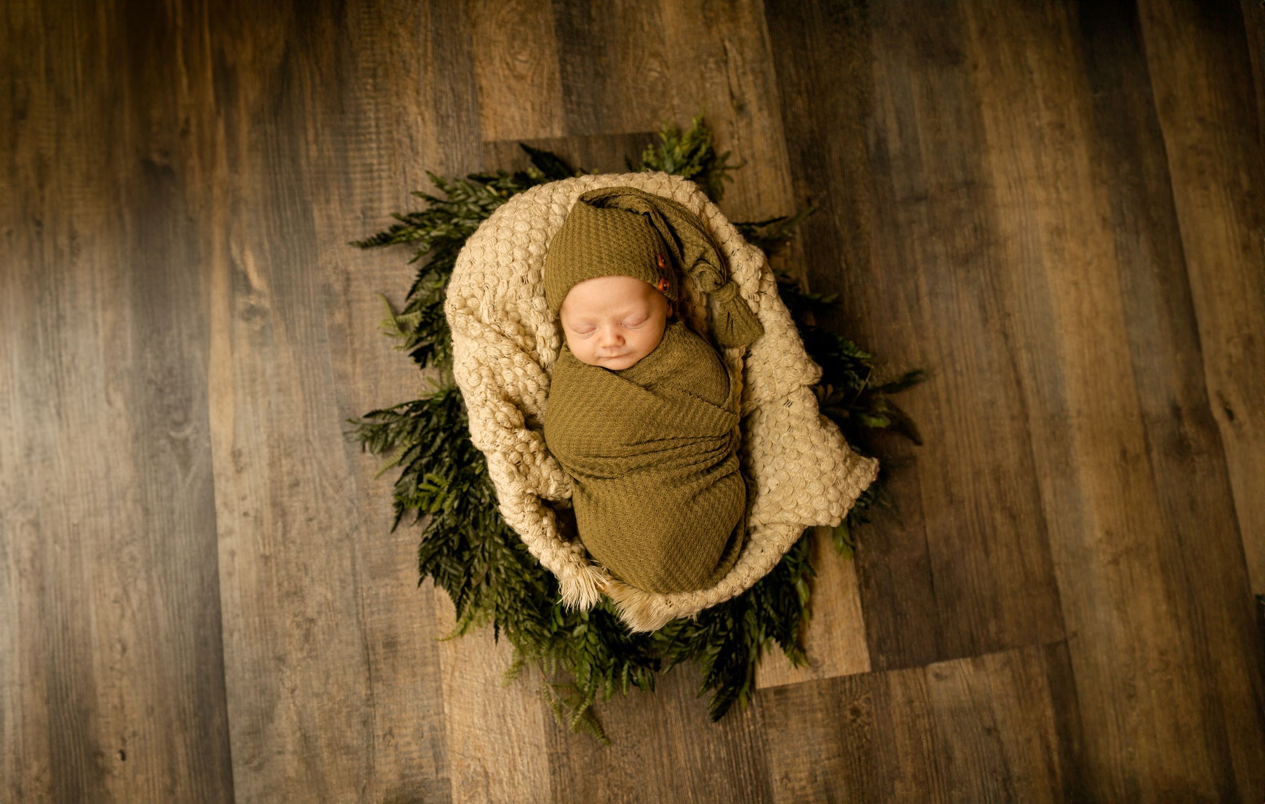 A sleeping newborn wrapped in beige fabric on a wreath of greenery against wooden flooring.