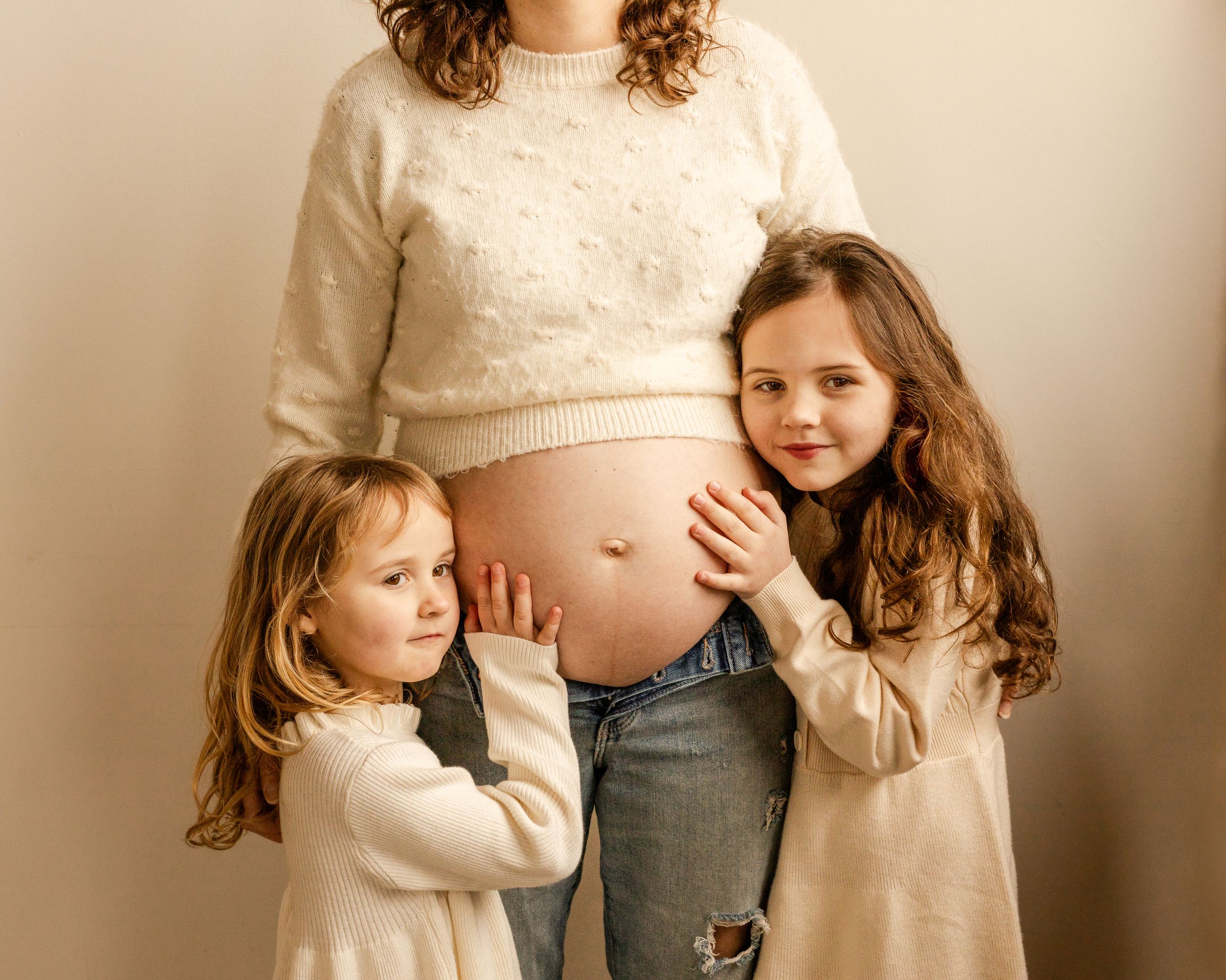Two children in cream sweaters embrace their mother's pregnant belly in a studio setting.