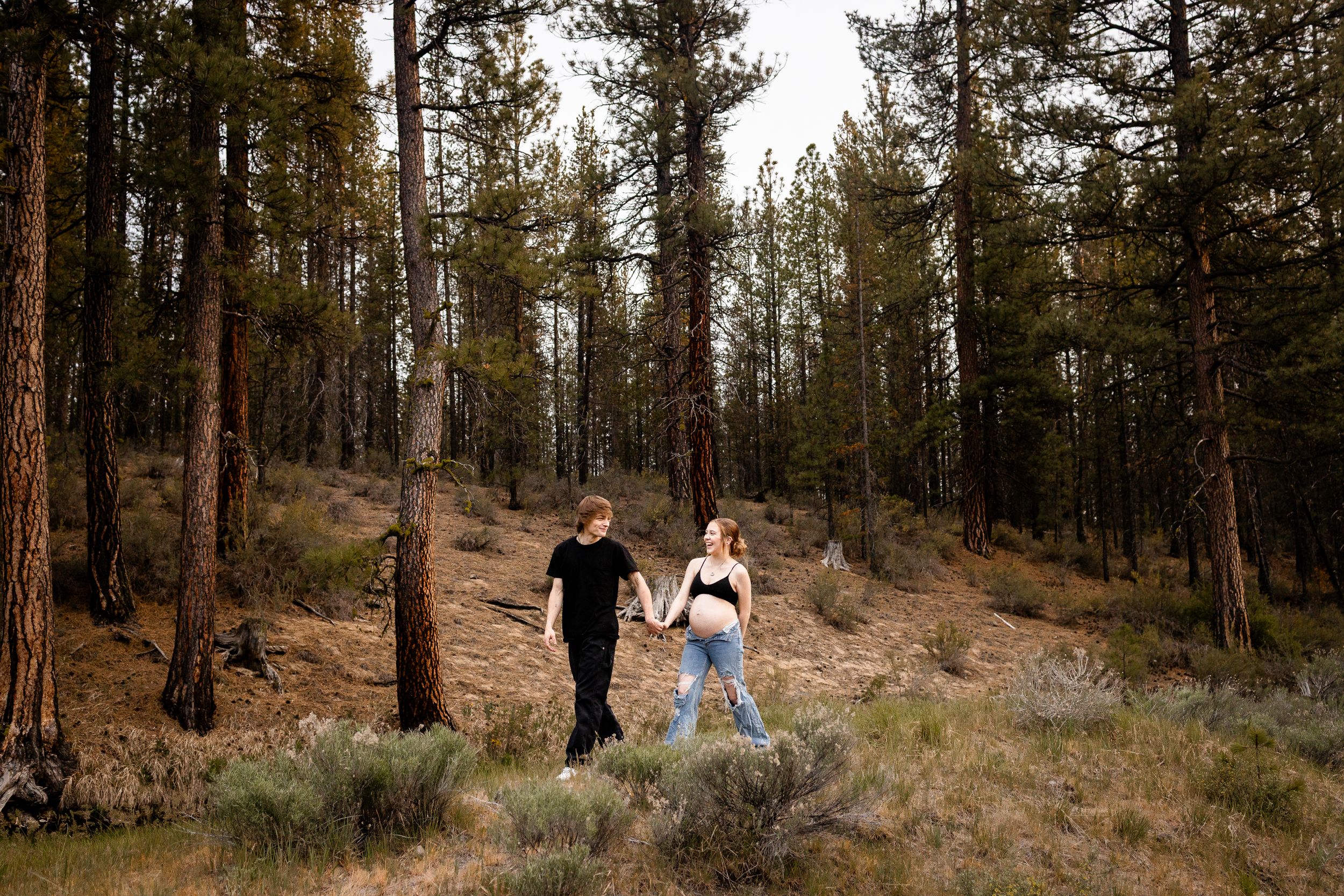 A couple walks hand in hand through a serene pine forest on a sloping hillside at sunset. Outdoor maternity photos in jeans.