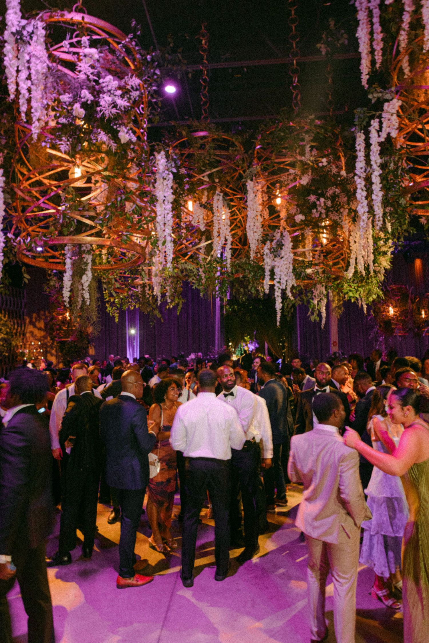 An elegant wedding venue with hanging floral decorations and purple lighting over a crowded dance floor.