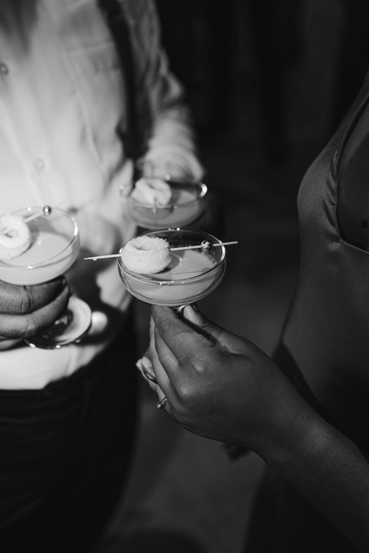 Black and white photo of hands holding garnished cocktail glasses at an event.