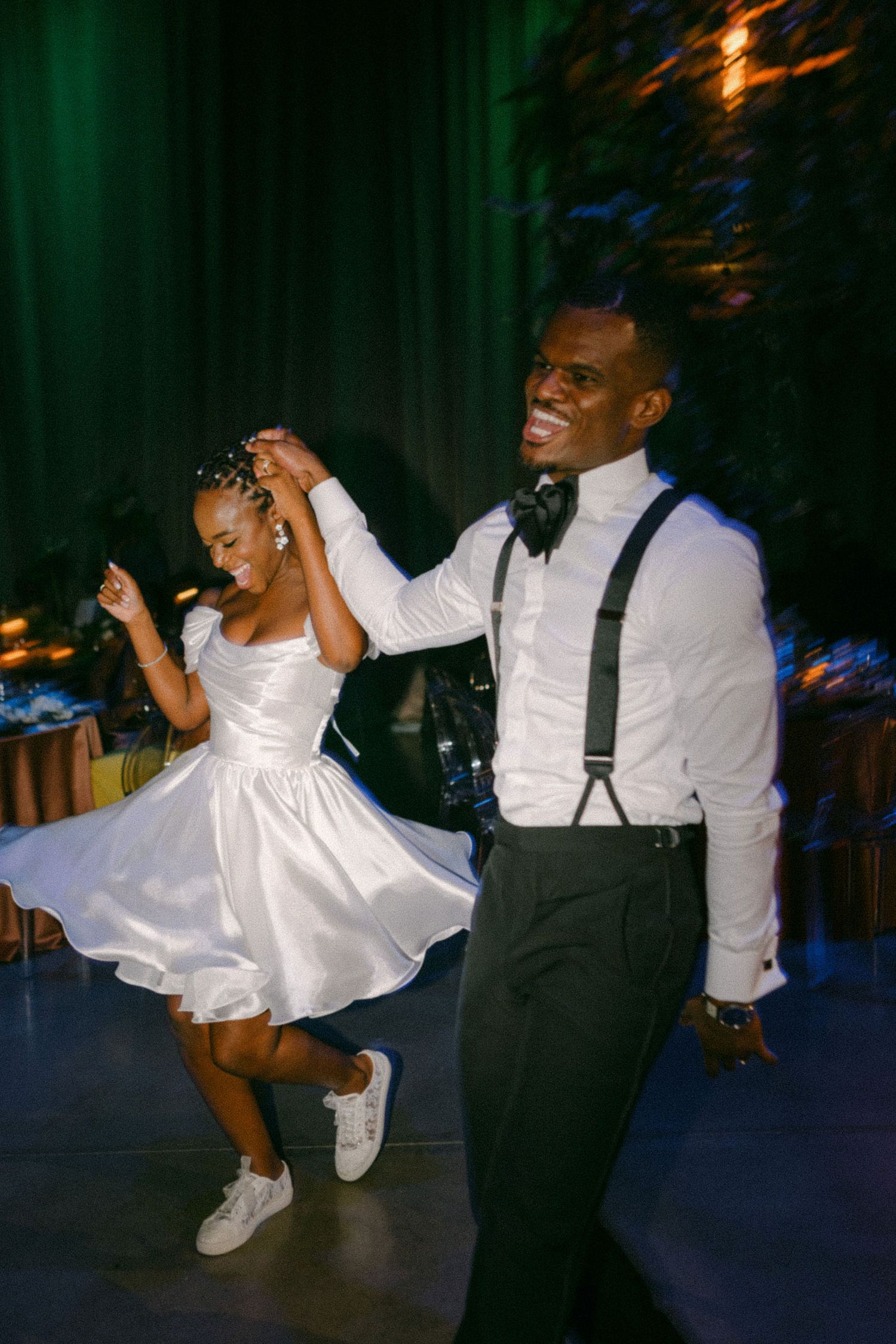 A couple dances energetically at a wedding reception, the bride wearing a short white dress and sneakers.