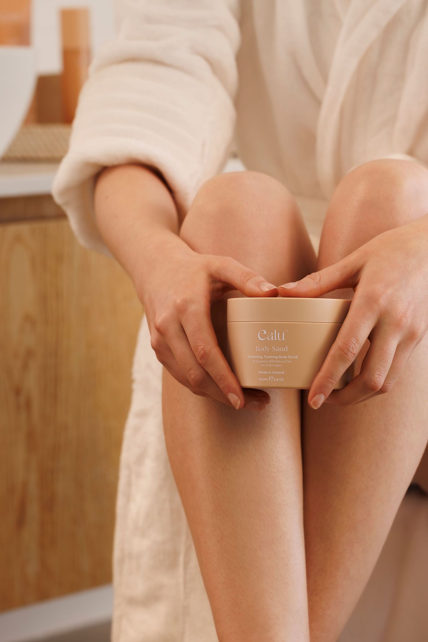 Hands holding skincare product jar while sitting in cozy white bathrobe.