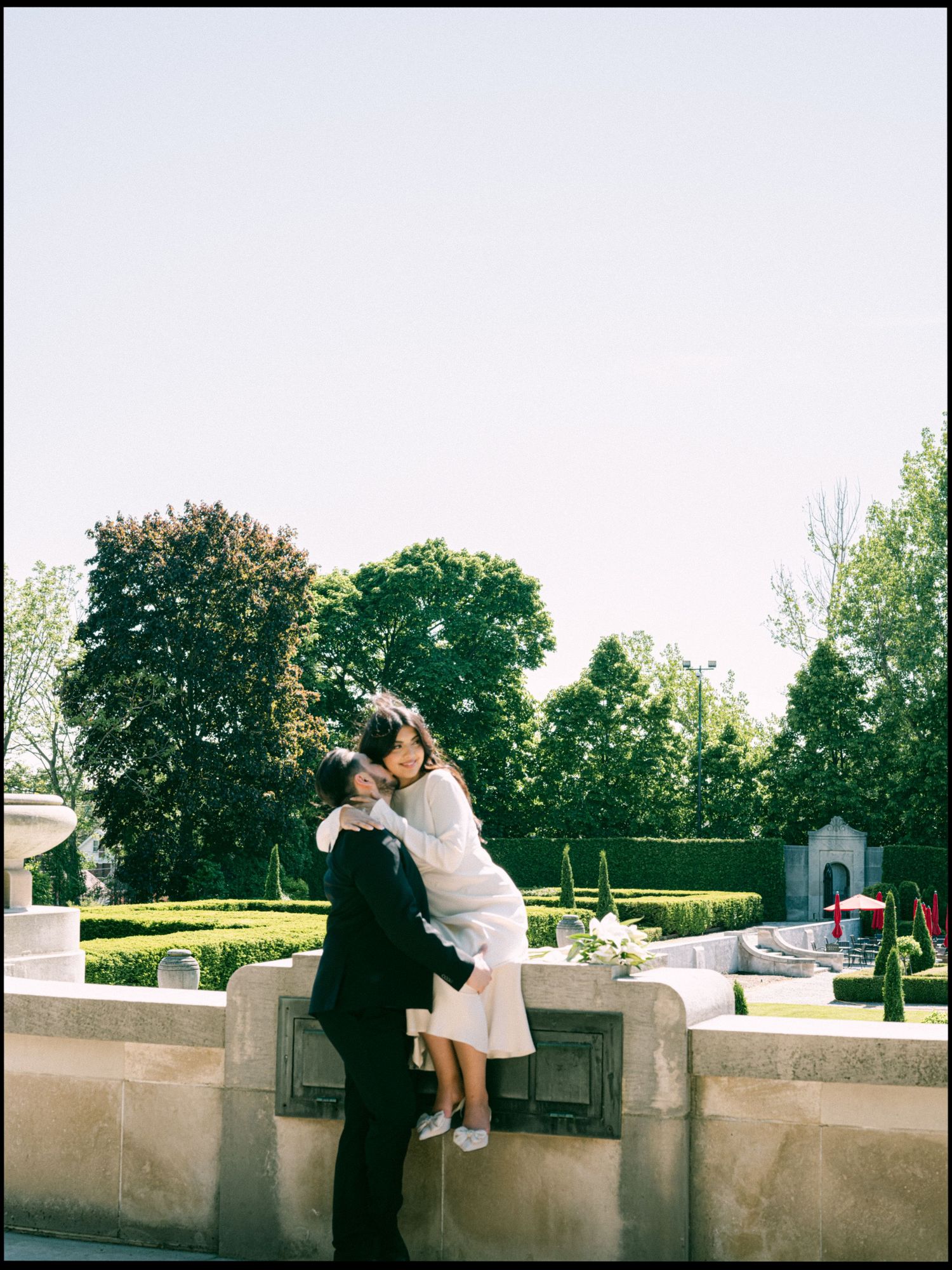A couple sharing a tender moment on a stone wall in a garden setting.
