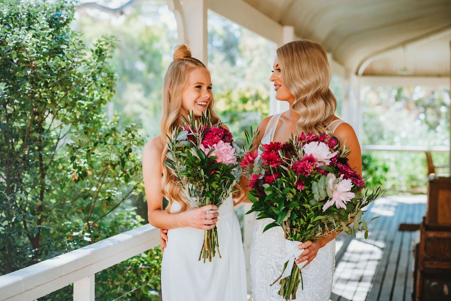 Two women in white dresses holding large bouquets of pink and white flowers on a covered porch.