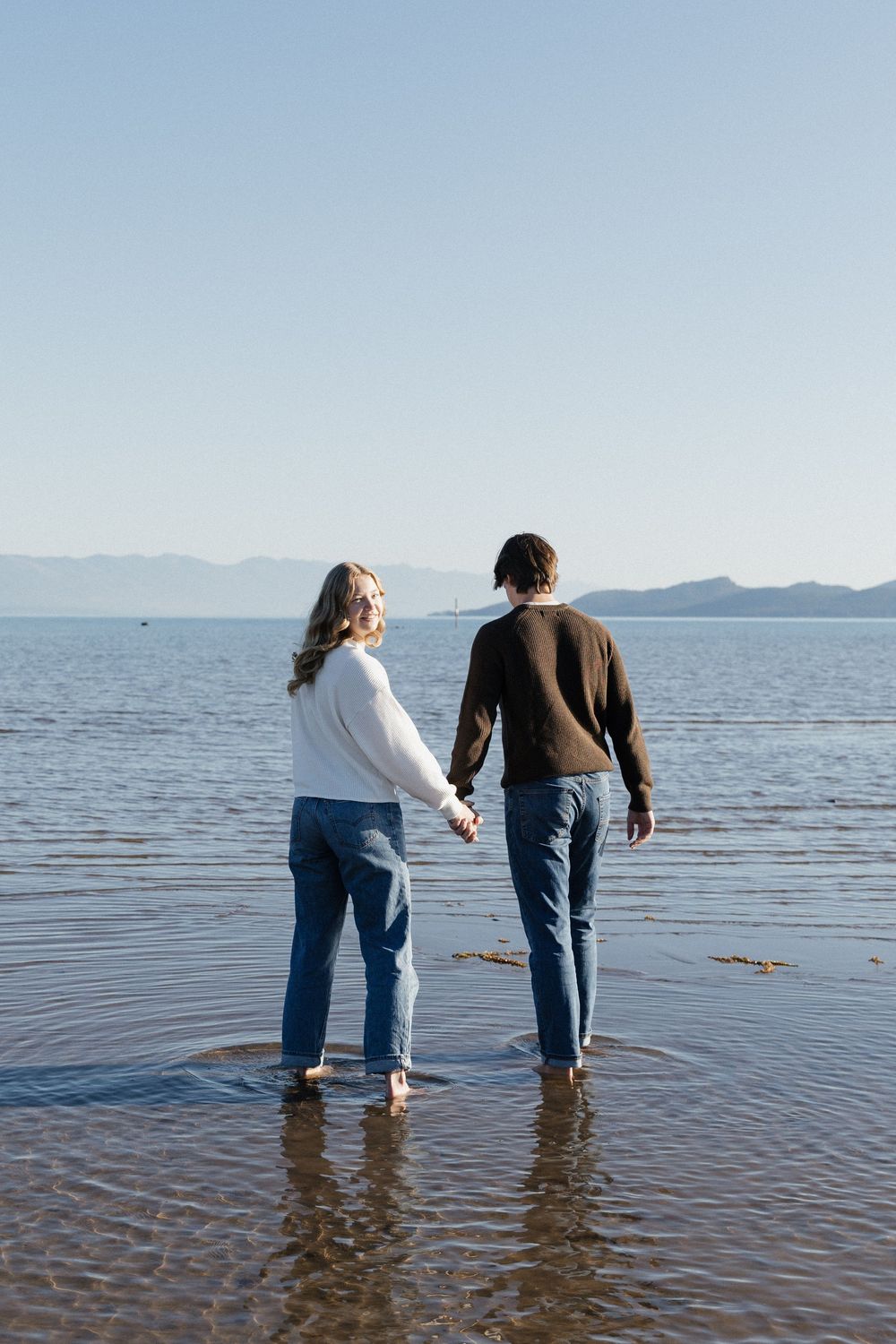 Two people wade together in shallow waters of a serene lake with mountains visible in the distance.
