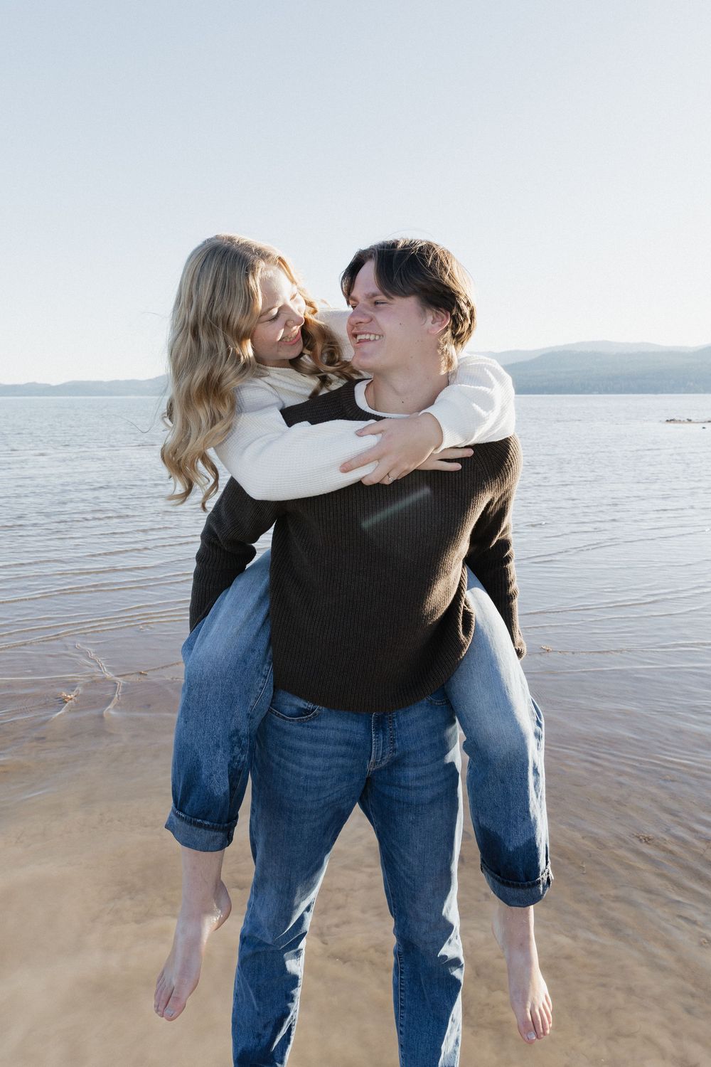 A couple embraces playfully on a sandy beach at sunset with the ocean in the background.