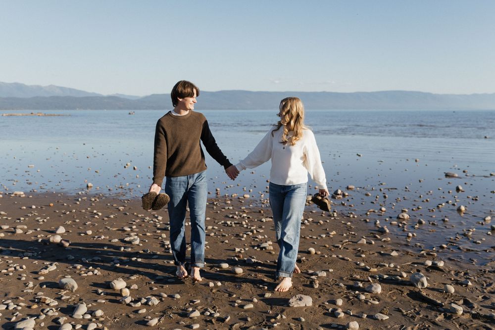 A couple walks hand in hand along a rocky beach shoreline with mountains visible in the distance.
