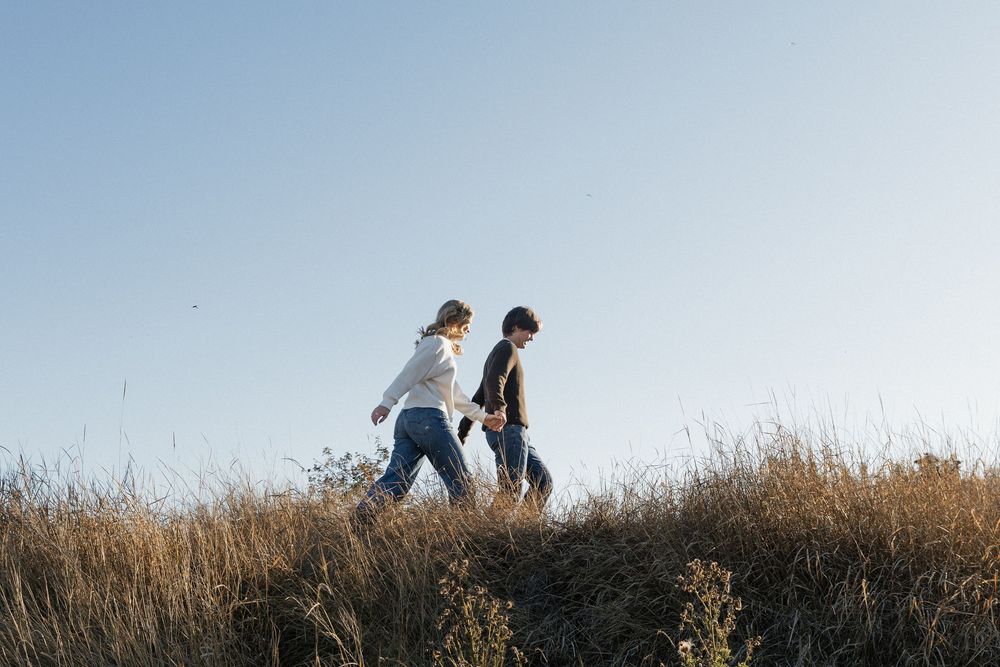 Two people walk together along a grassy hilltop silhouetted against a bright cloudy sky at sunset.