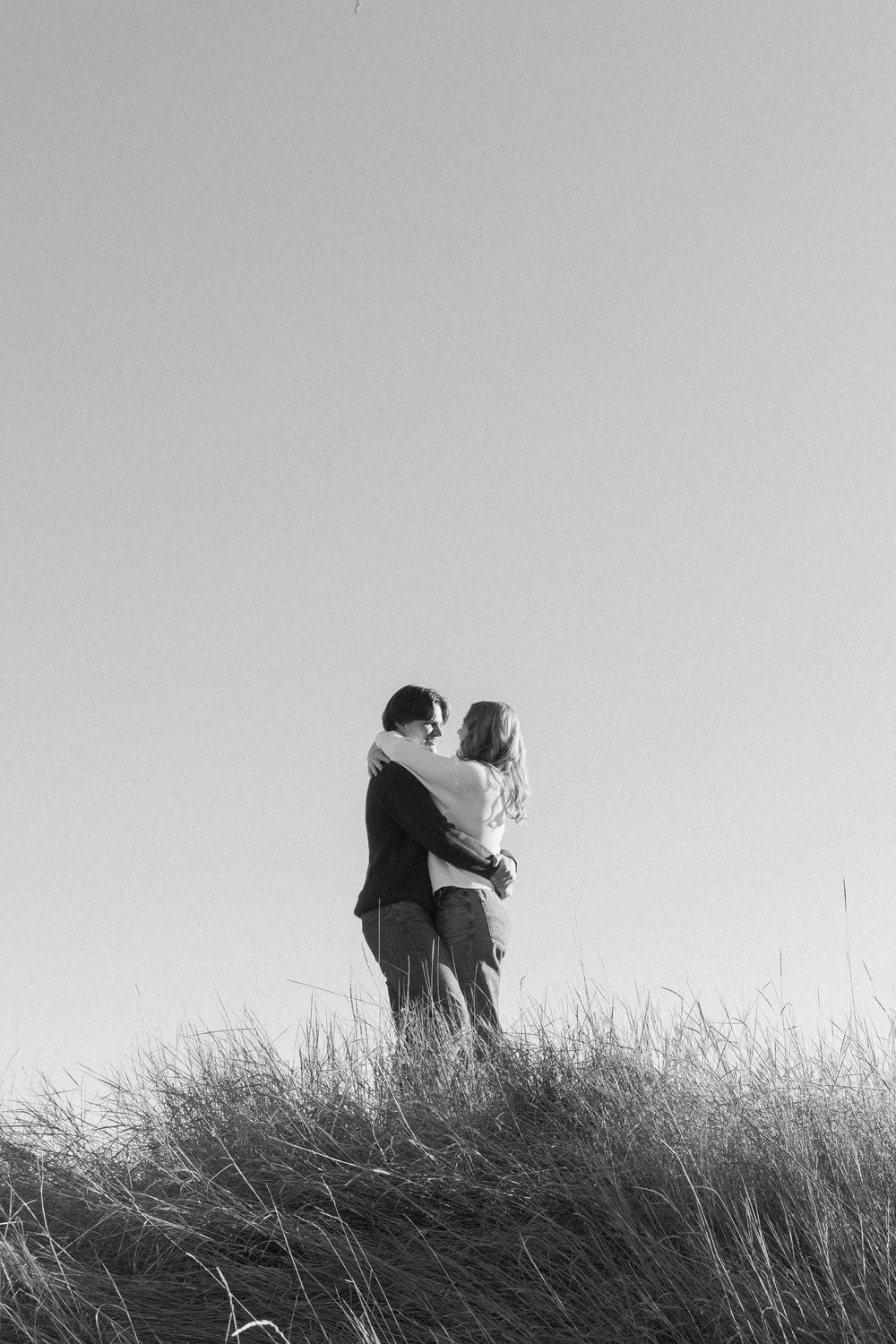 A black and white silhouette shows two figures embracing on a grassy hilltop against a stark white sky.