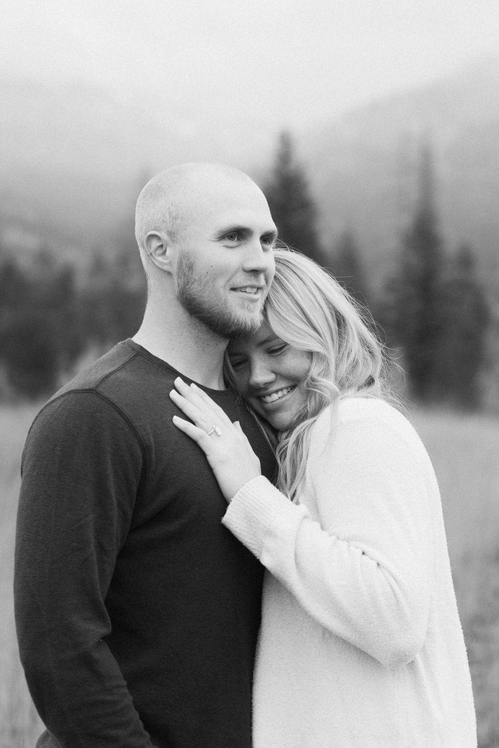 A romantic black and white engagement photo of a couple embracing outdoors with mountains and evergreens in background.
