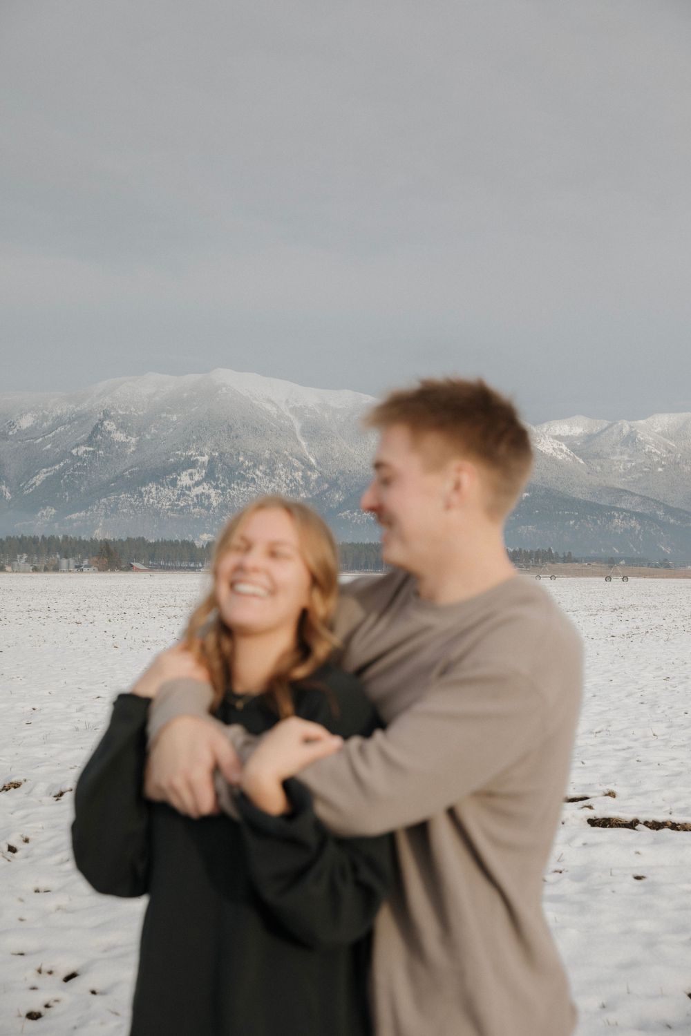 Couple embracing and laughing in front of snow-capped mountains on a winter day.