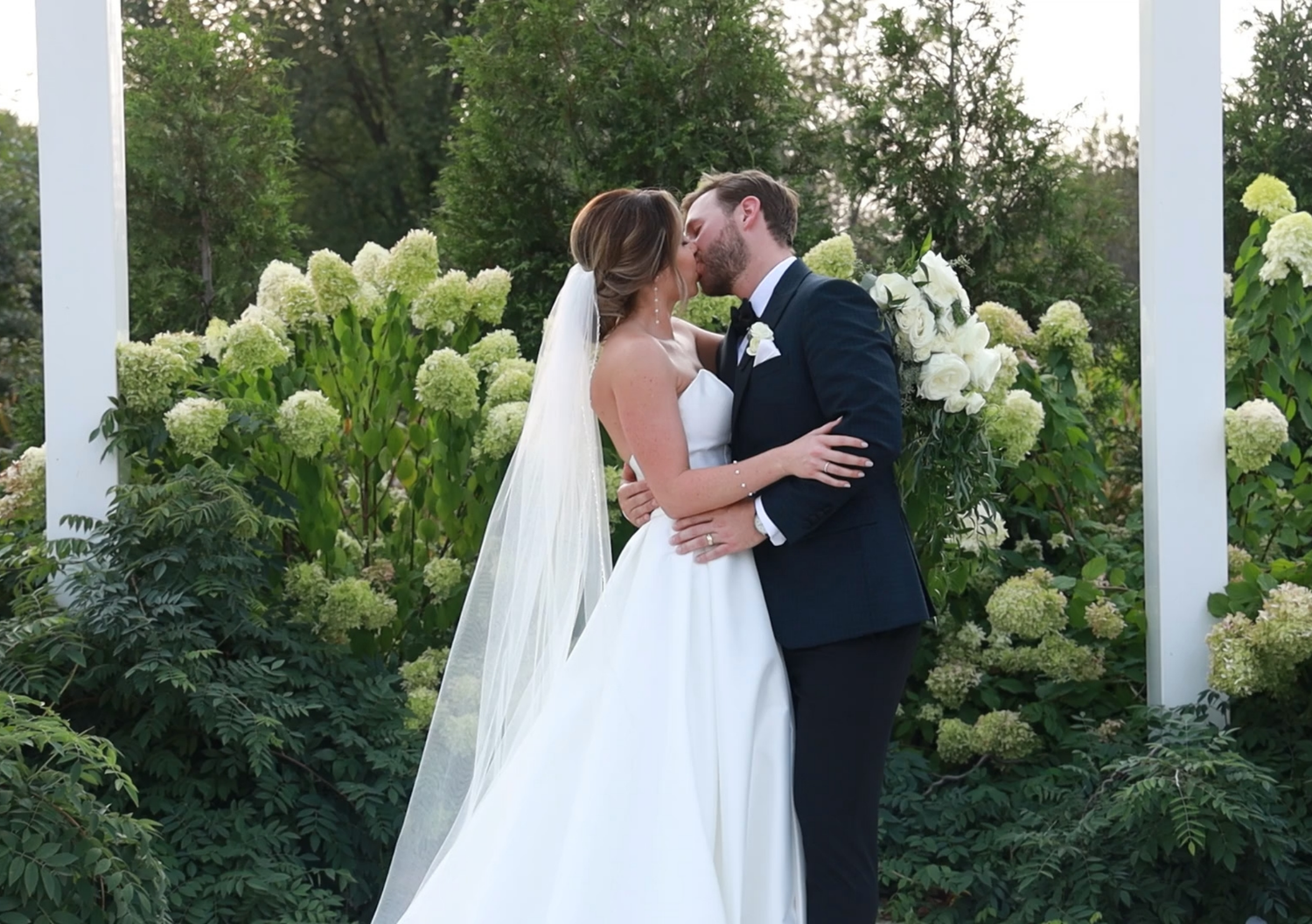 A tender wedding kiss captured in a garden setting with blooming white hydrangeas and elegant columns.