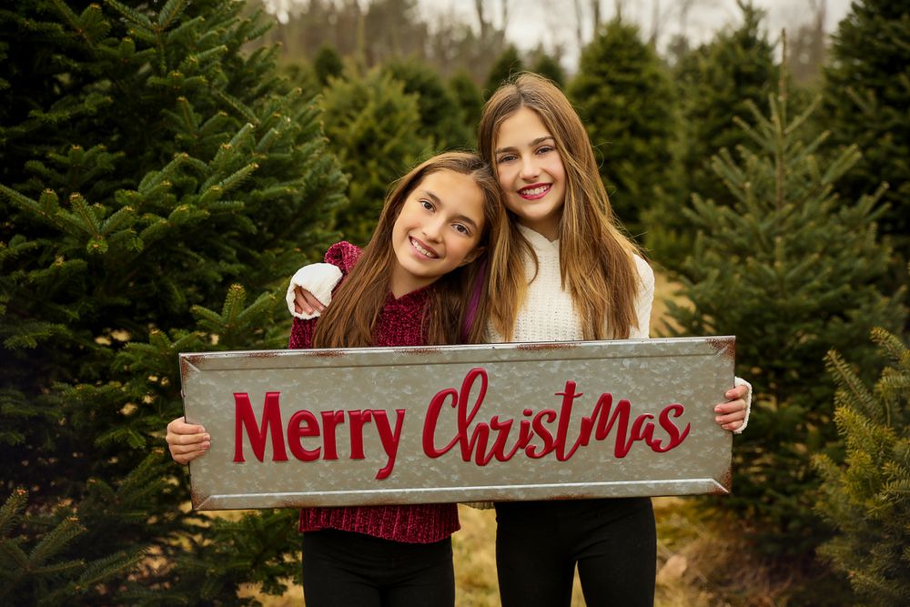 Two teens in a Christmas tree farm holding a merry Christmas sign