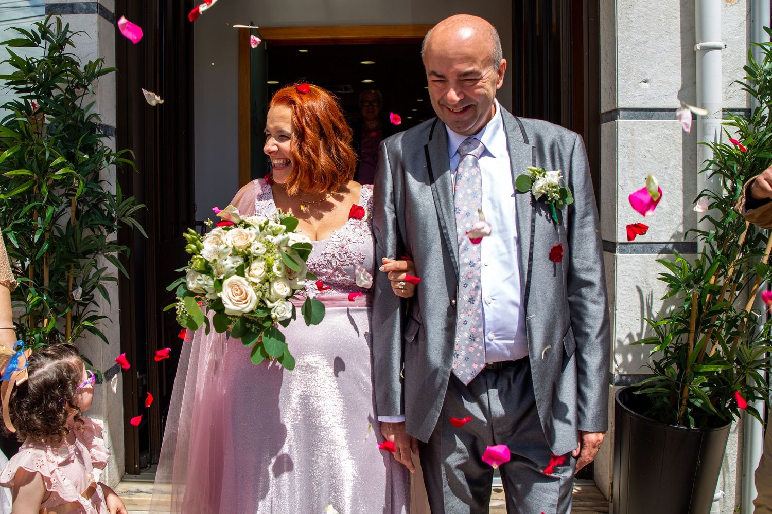 A wedding couple exits their ceremony venue as rose petals fall around them.