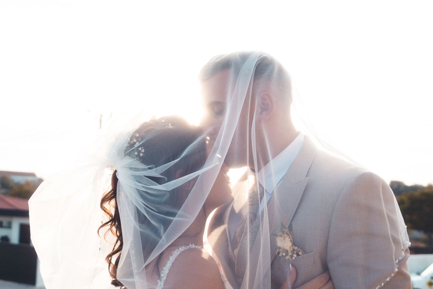 A romantic wedding couple shares a kiss under a delicate veil with sunlight streaming through.