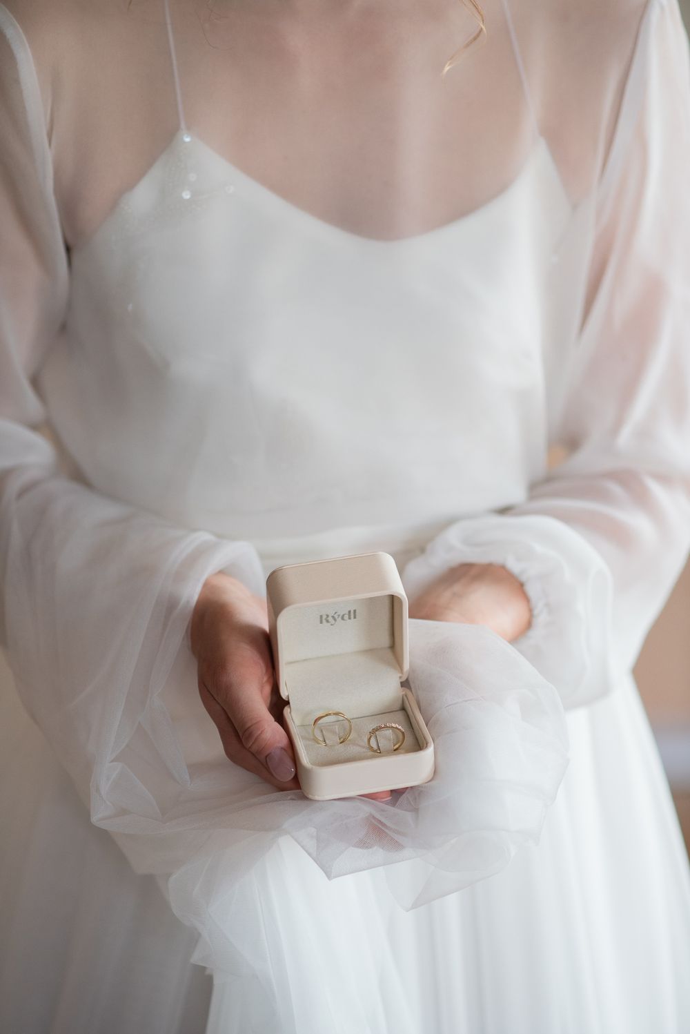Wedding rings in small white box held by person in flowing white dress.