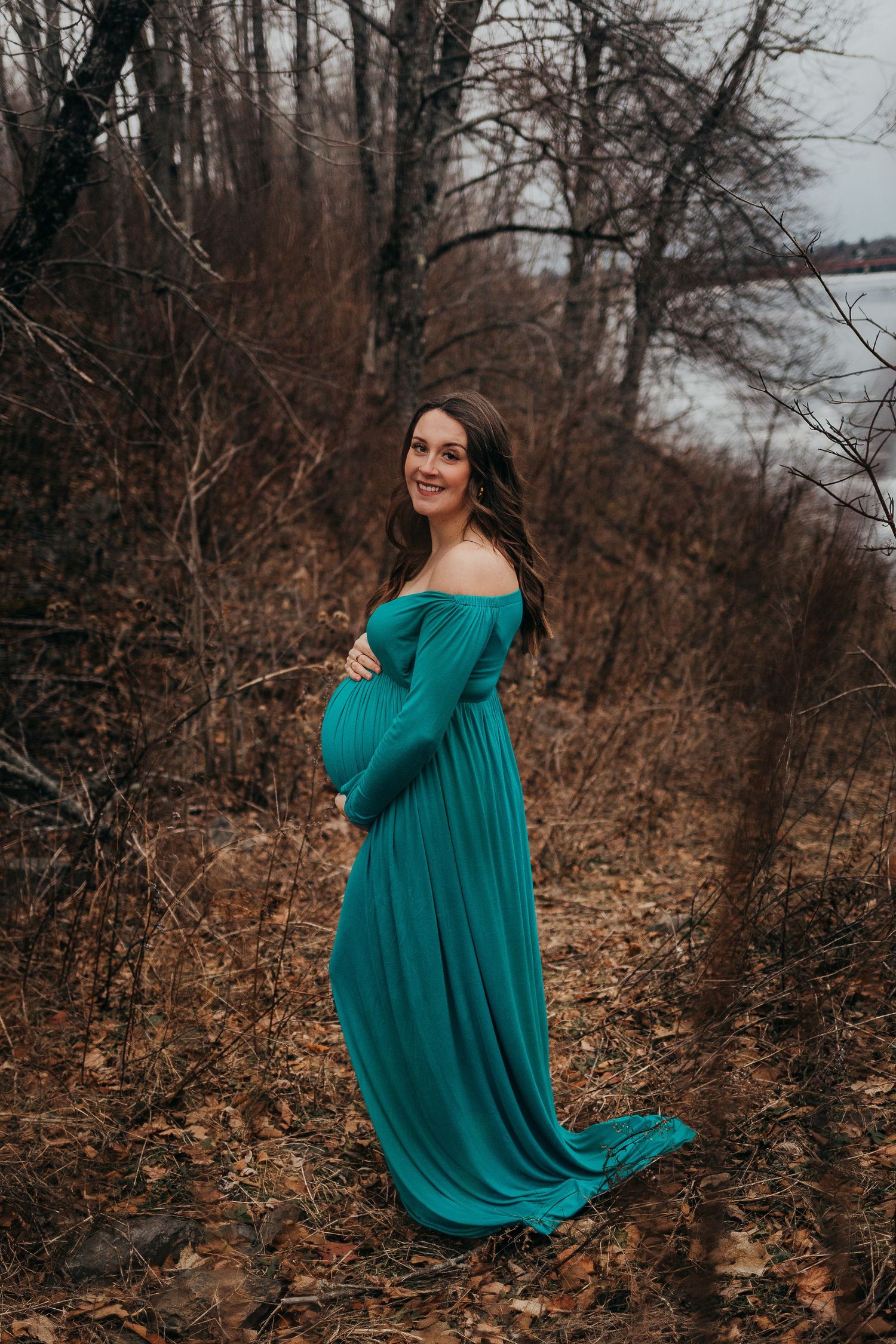 A woman wearing a flowing teal off-shoulder maternity gown poses in an autumn forest setting.