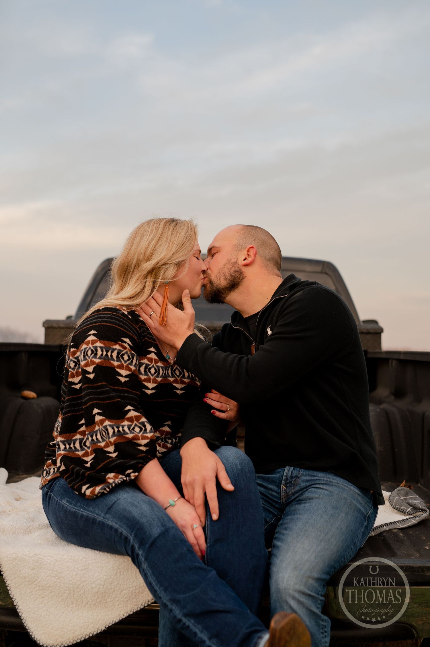 A romantic couple kisses while sitting on a truck bed with a blanket during sunset.