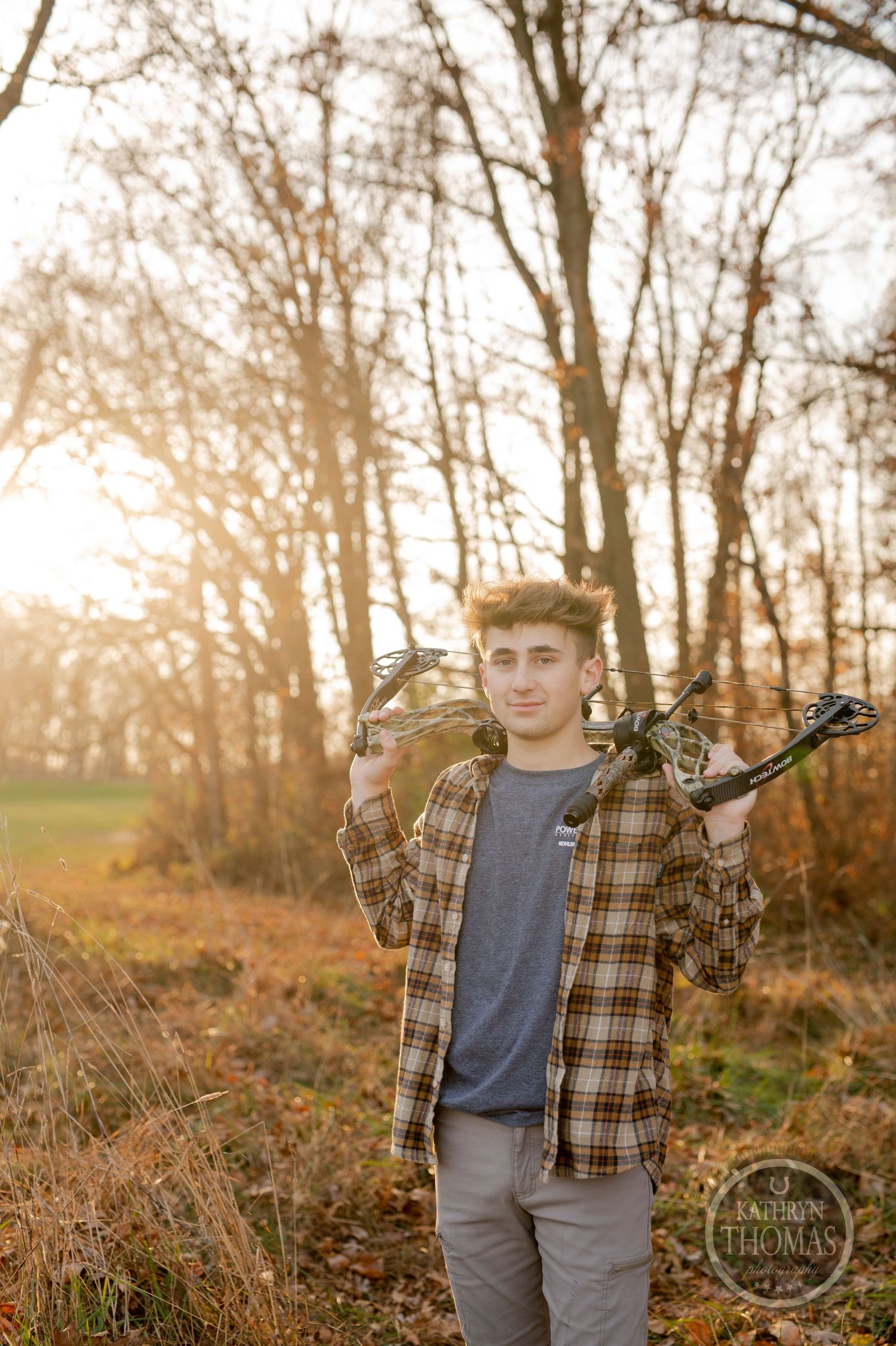 Hunter carries compound bow across shoulders while standing in autumn woods at sunset.