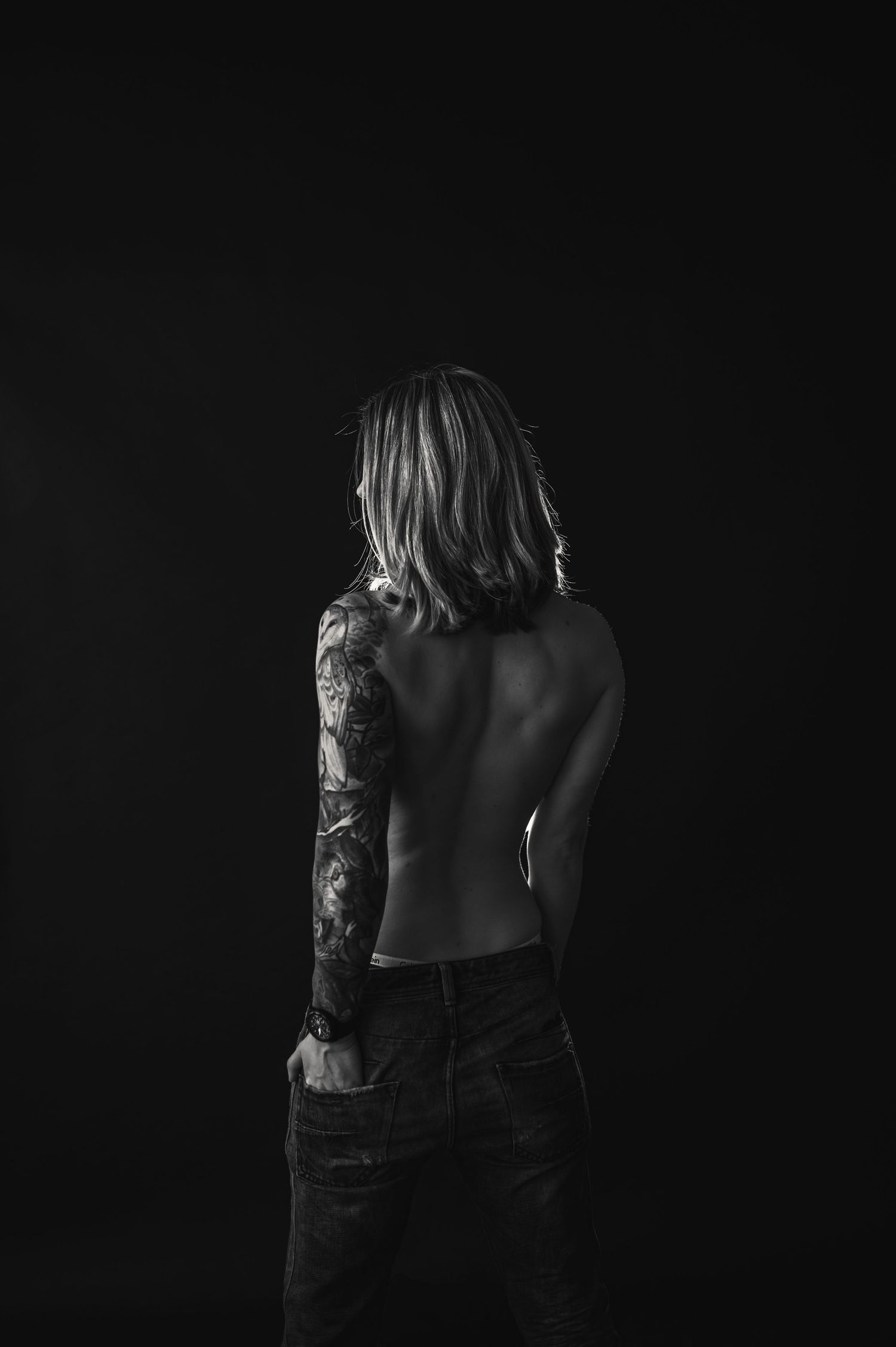 Artistic black and white portrait from behind showing a bare back and ornate sleeve tattoo against dark background Boudoir West Michigan